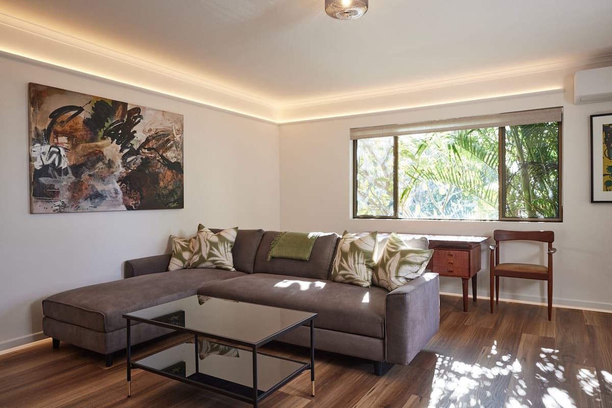 A modern living room with a sectional sofa, a glass coffee table, a wooden desk, and a large window with a view of greenery.