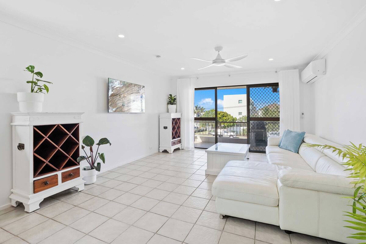 A spacious living room with a white sofa, a fireplace with a diamond-patterned cabinet, and a view through large windows.