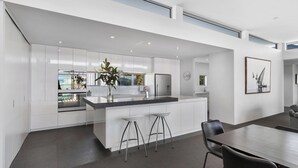 A modern kitchen with white cabinetry, a central island with stools, and a dining area with a table and chairs. - Lake & Mountain views, close to ski fields. Executive living - 3 bed/3 bath, spa (Wānaka)