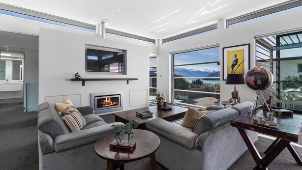A modern living room with a fireplace, grey sofas, a coffee table, and a large window offering a mountain view. - Lake & Mountain views, close to ski fields. Executive living - 3 bed/3 bath, spa (Wānaka)