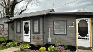 Ein eingeschossiges Haus mit grauer Fassade, einer Veranda mit Bank und einem kleinen Garten mit verschiedenen Pflanzen.