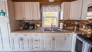 A kitchen with white cabinets, a double sink, a coffee maker, and a window with a view of trees. - Pelicans View - Venture Out Lot 318 (Cudjoe Key)