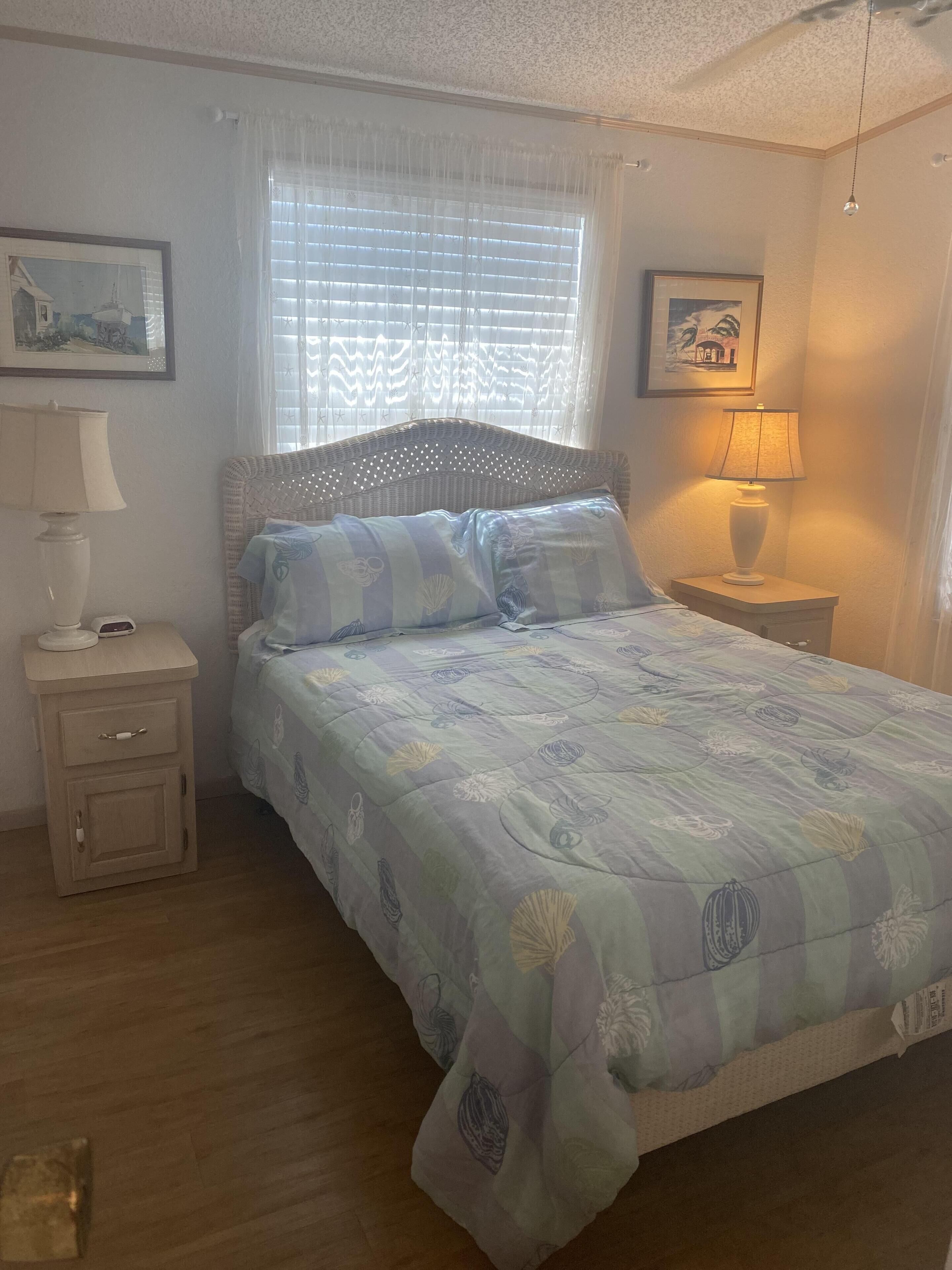 Une chambre à coucher comprenant un lit, une table de chevet, une lampe, une fenêtre avec des stores et un tableau accroché au mur.