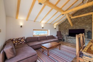 Un salón estilo loft con techo de madera, chimenea de piedra, un ventanal grande y un sofá de cuero marrón.