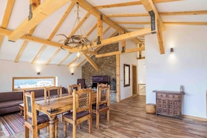 Una habitación amplia con techo de madera alto, una mesa de comedor con sillas, una chimenea de piedra y un ventanal grande.