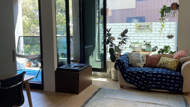 A modern living room with a sofa, a dining table, and a chair. There is a glass door leading to an outdoor area with plants and a sign that reads "IMAGINE EQUALITY".