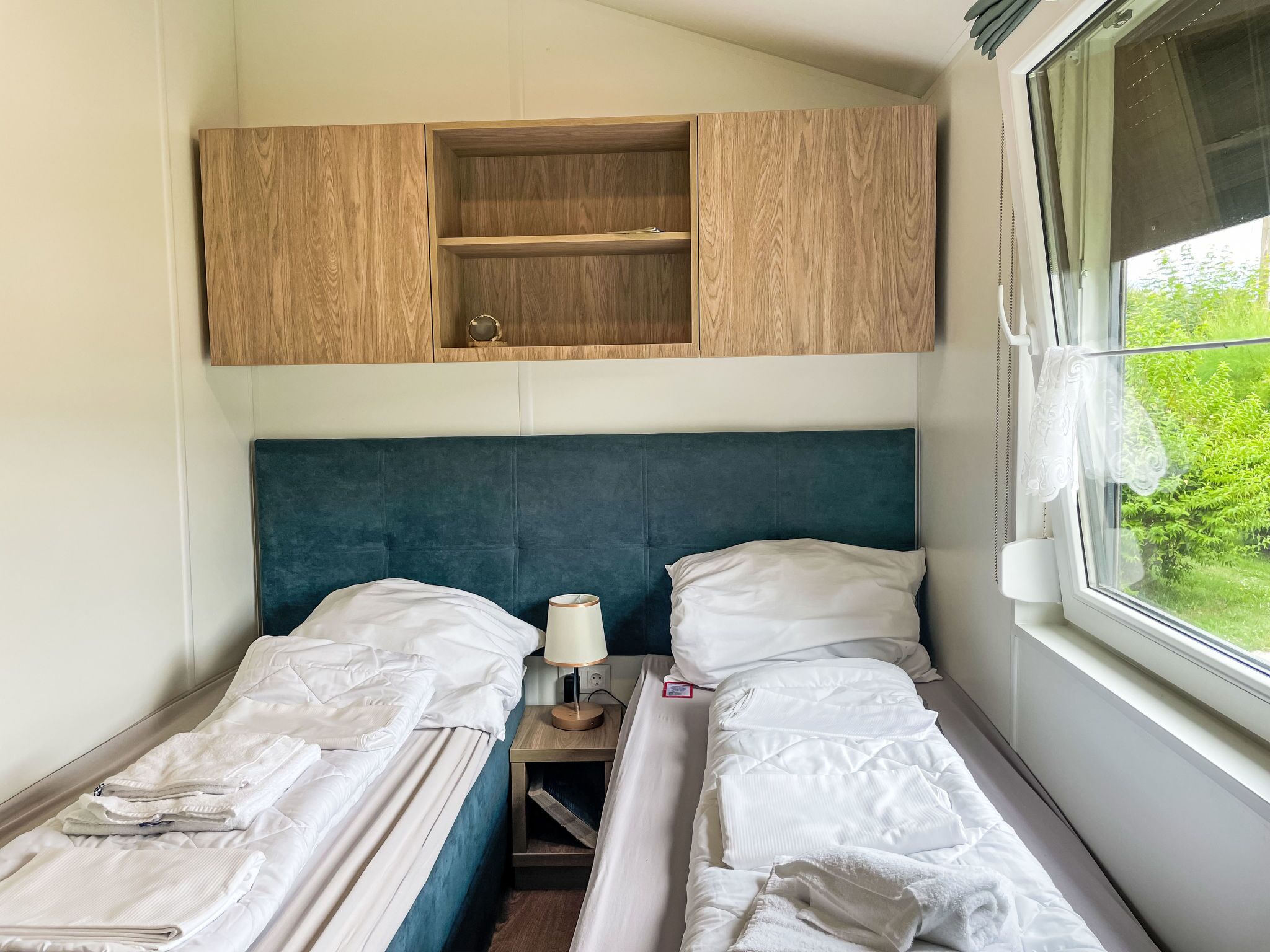 Une petite chambre, de taille modeste, avec deux lits simples, un placard en bois au-dessus et une fenêtre donnant sur un paysage verdoyant.