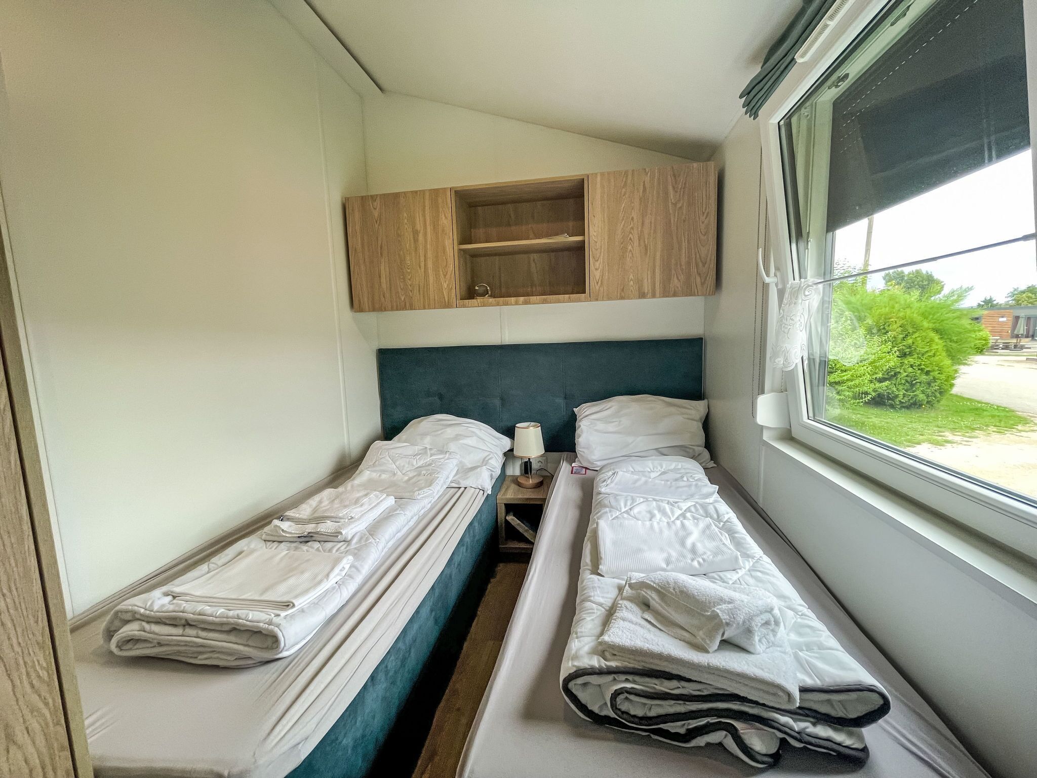 Een kleine kamer met twee bedden, een houten plank erboven, een bedlampje en een raam met uitzicht op groen.