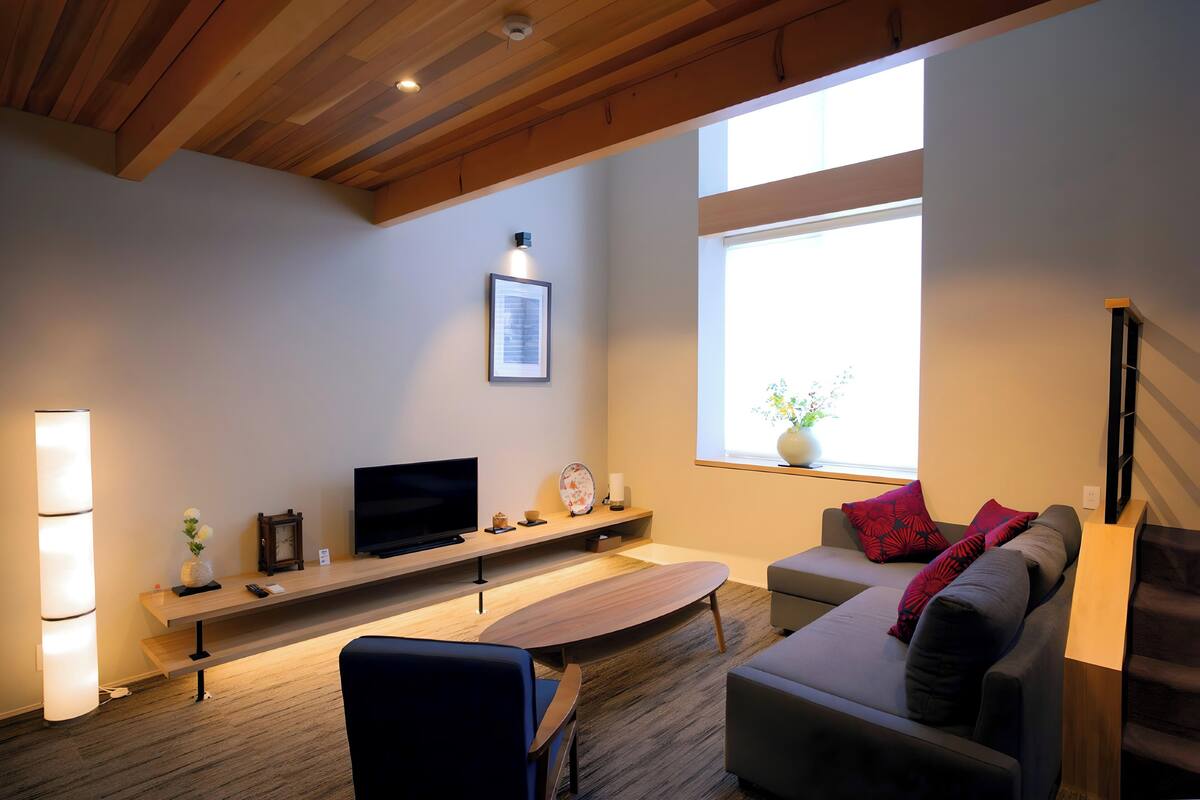 A modern living room with a wooden ceiling, a flat-screen TV, a wooden coffee table, a sofa, and a window with a view of greenery.