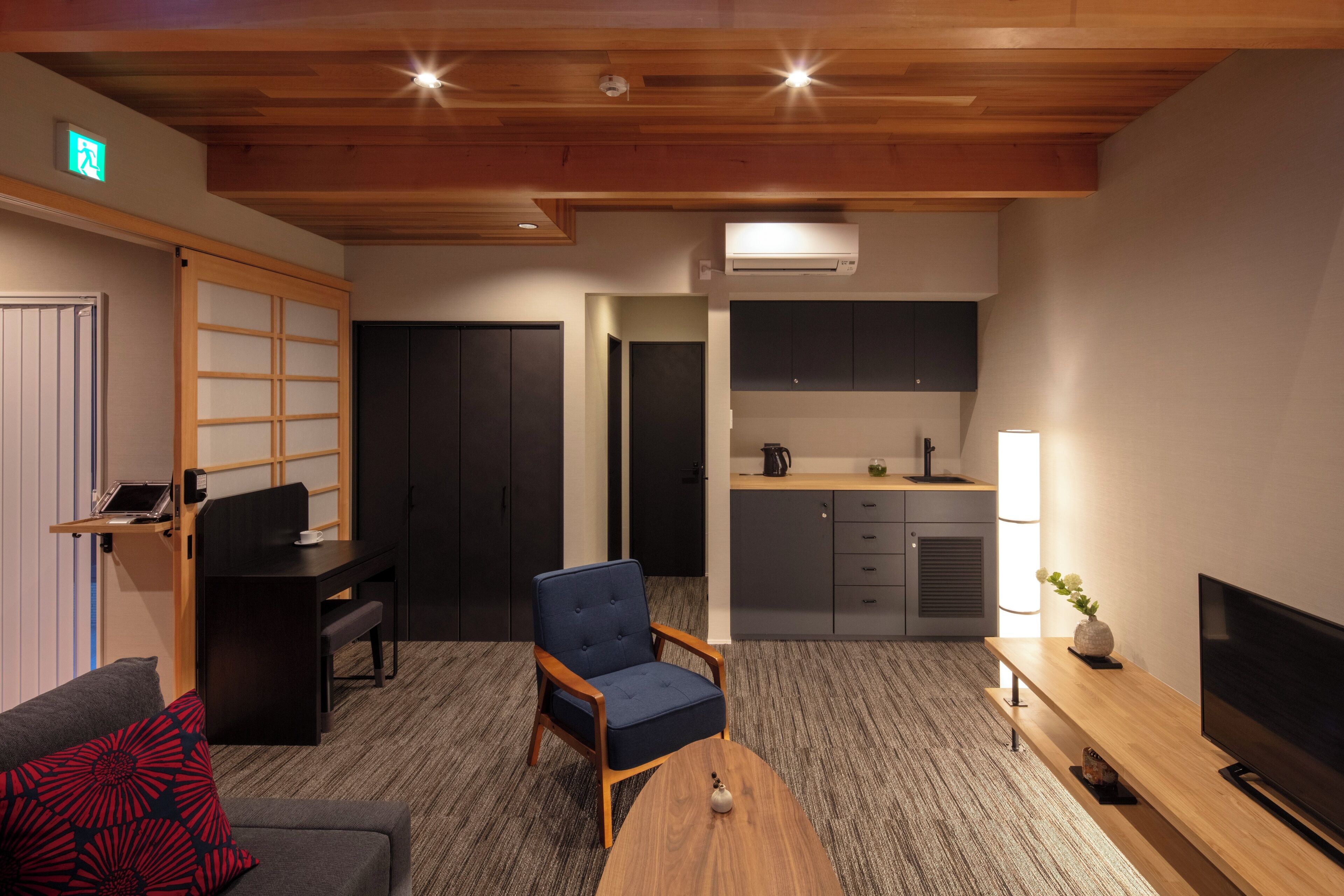 Una habitación de hotel moderna con techo de madera, un sofá con almohada roja, un sillón azul, una mesa de centro de madera, un televisor sobre un mueble de madera, una zona de cocina con armarios y una tetera, y una planta pequeña sobre una mesita.