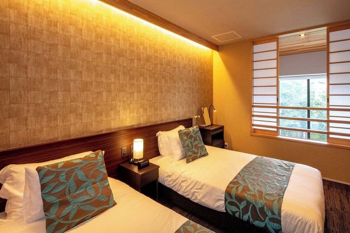 A hotel room with two beds, a nightstand with a lamp, a window with blinds, and a view of greenery outside.