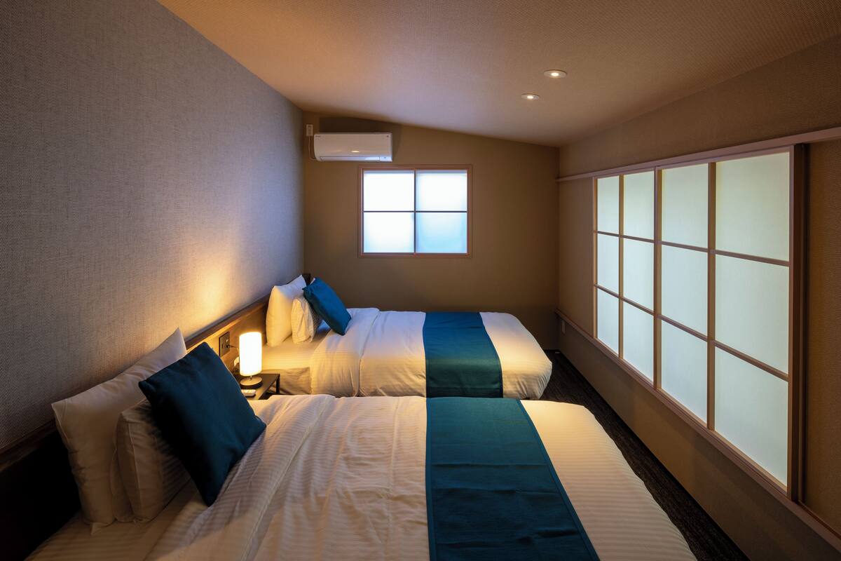 A hotel room with two beds, a window with frosted glass, and a wall-mounted air conditioner.