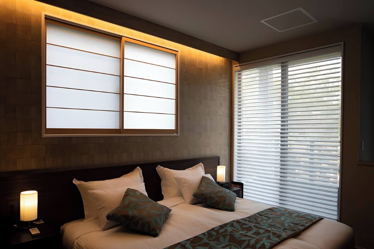 A modern hotel room with a large bed, bedside lamps, a window with blinds, and recessed ceiling lighting.
