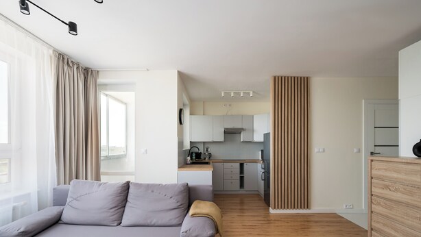Een moderne woonkamer met een grijze bank, houten vloer en een keuken op de achtergrond.