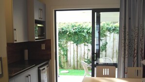 Una cocina compacta equipada con lavavajillas, microondas y un mueble pequeño. Hay una zona de comedor con mesa y sillas, y un ventanal corredero que da a un patio trasero con cerca de madera y vegetación.