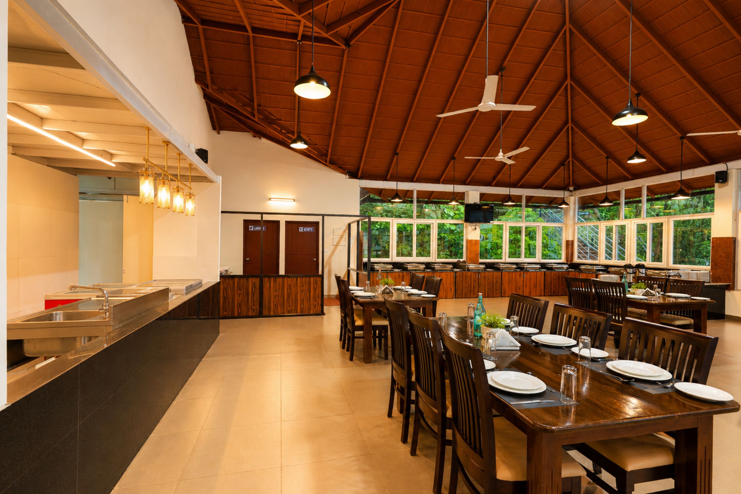 A spacious dining area with a high ceiling, multiple tables, and chairs, a buffet counter, and large windows offering a view of greenery.