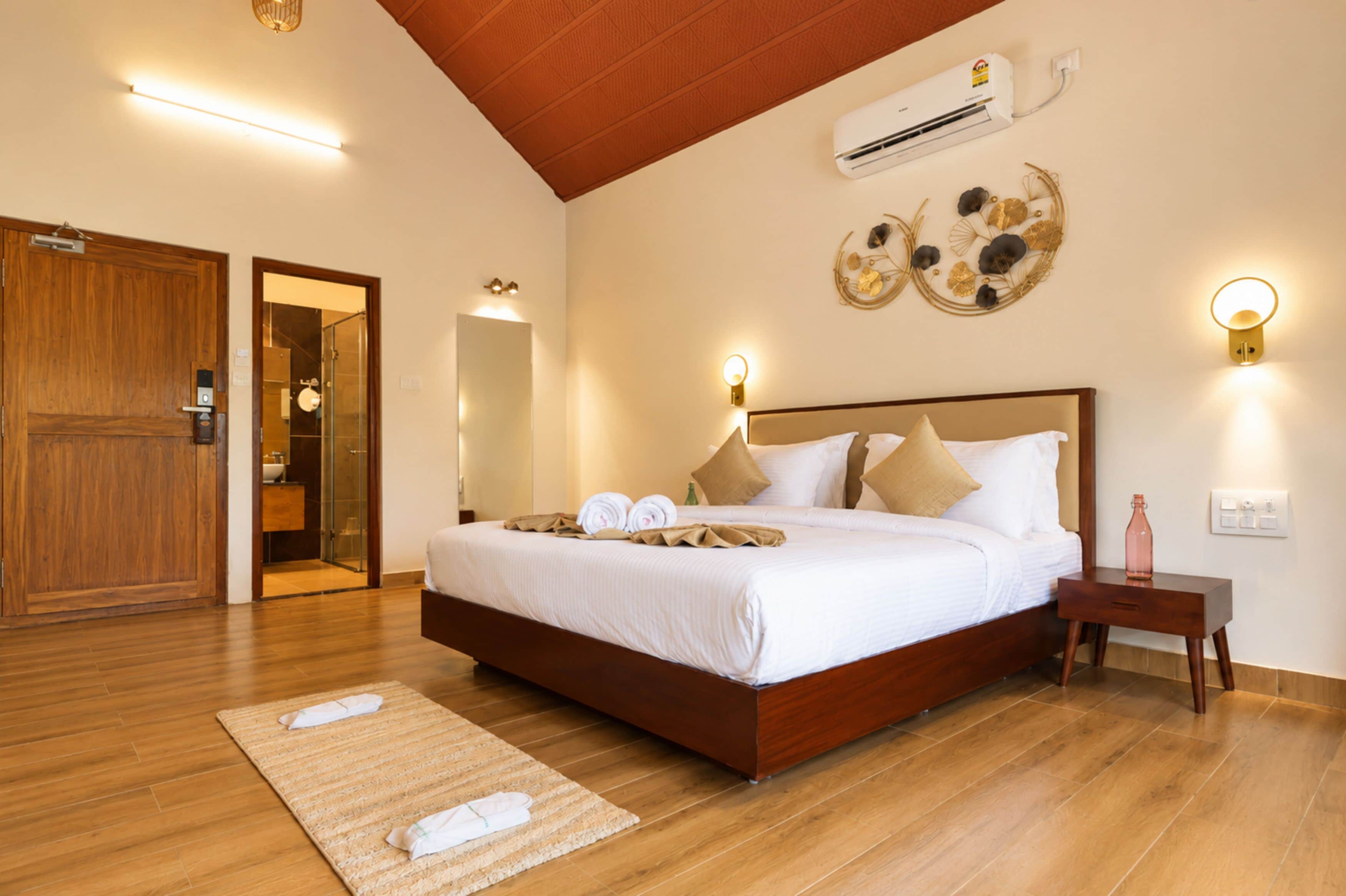 A hotel room with a wooden floor, a bed with white bedding and two pillows, a nightstand with a bottle, and an air conditioning unit on the wall.