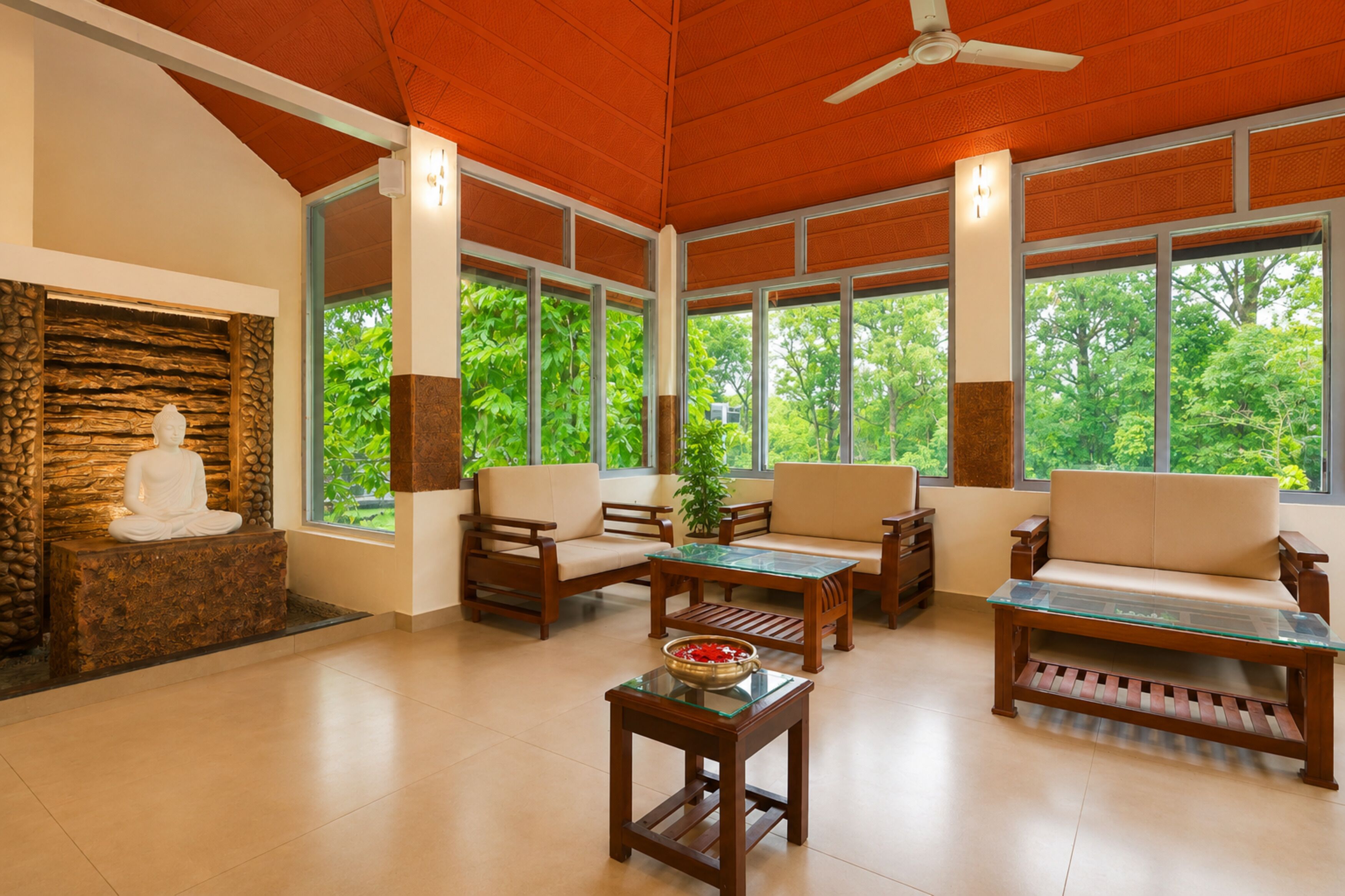 A spacious room with a stone wall, a Buddha statue, wooden furniture, and large windows with a view of greenery.