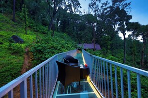 A glass bridge with wicker furniture and a table, surrounded by lush greenery and trees. - Silent Brook Resort - Coorg (Madikeri)