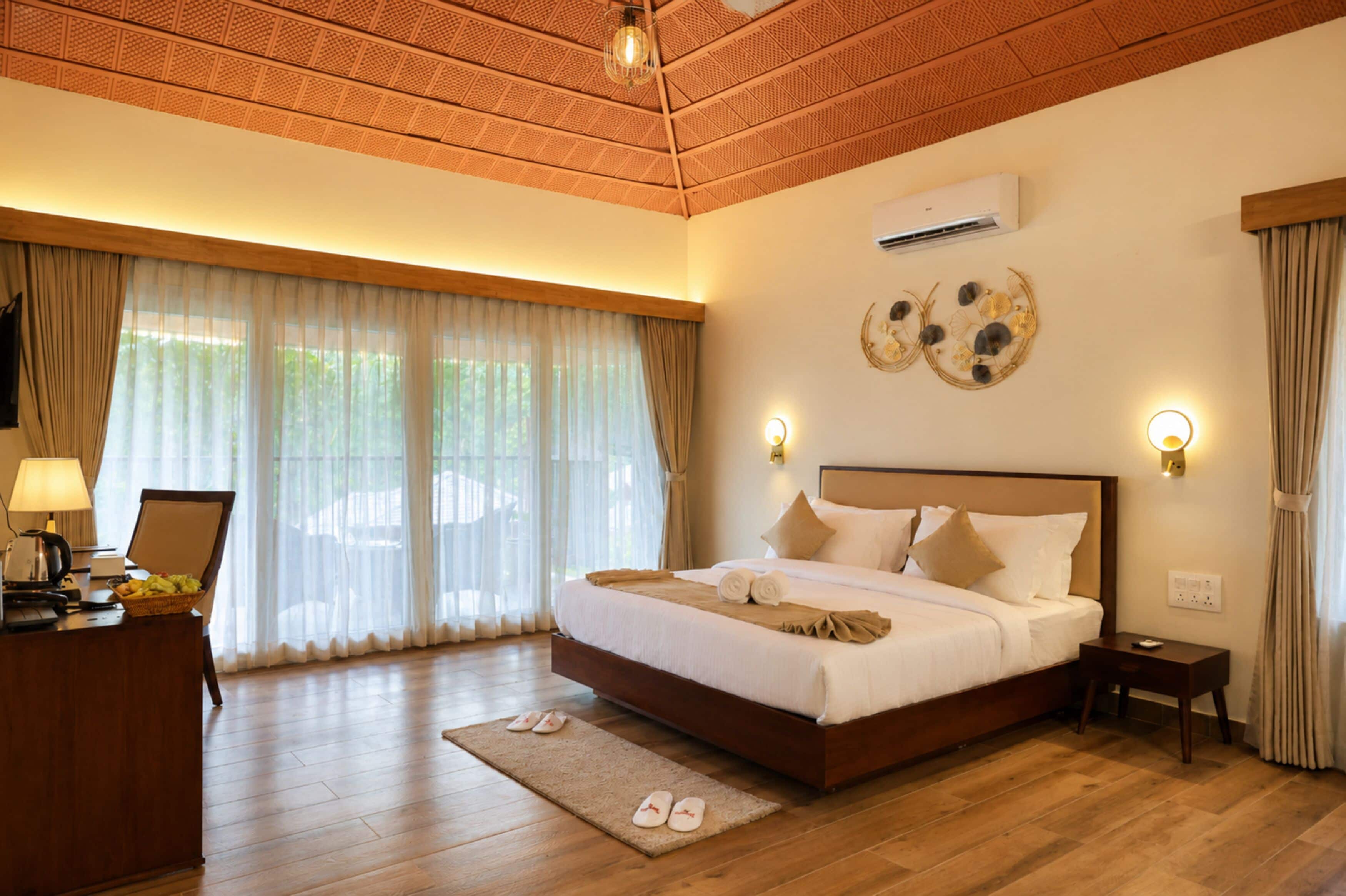 A hotel room with a large bed, a desk with a lamp, and a view of greenery through the window.