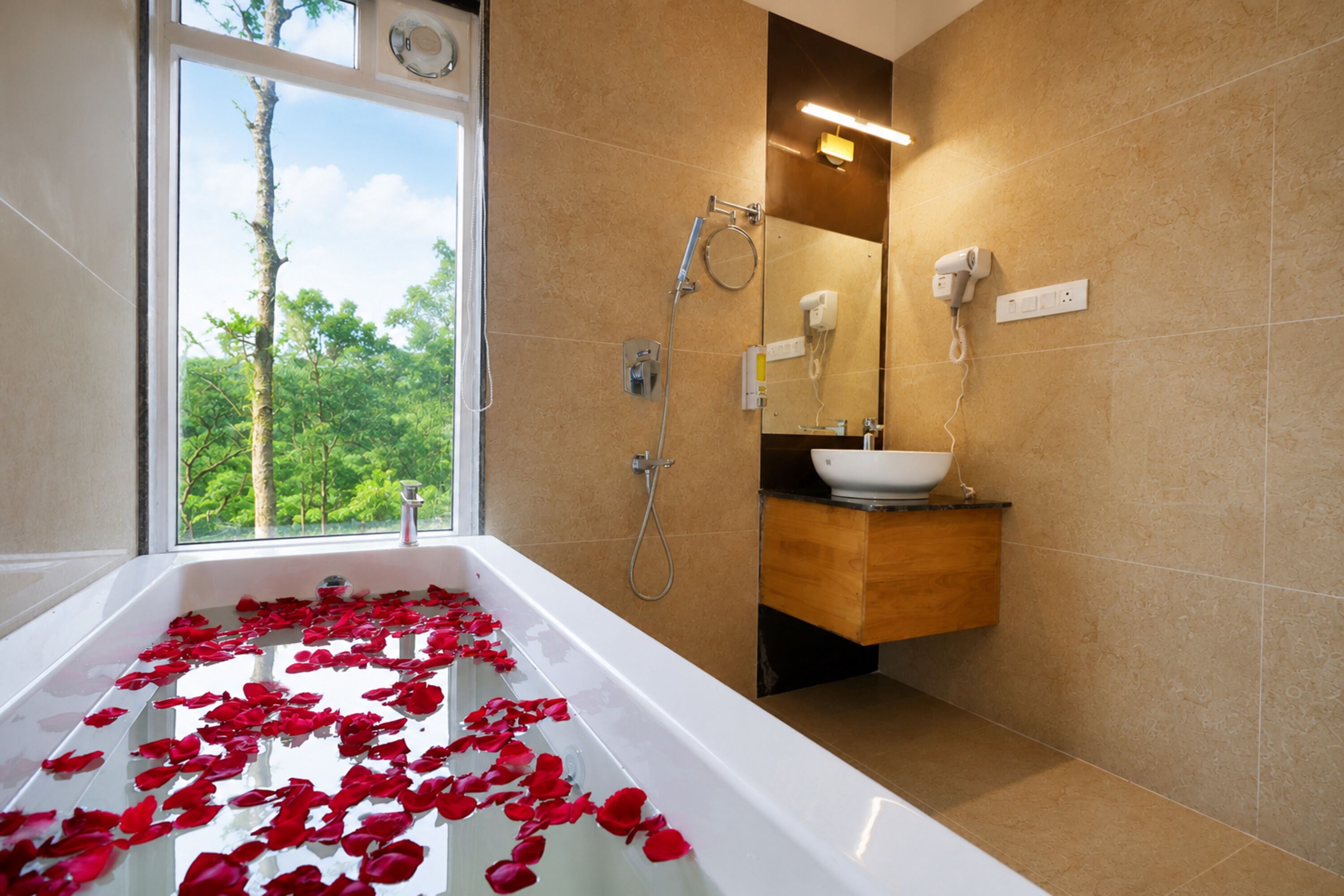 Un baño con una bañera llena de pétalos de rosa, una ventana con vistas a árboles y un tocador moderno con espejo.