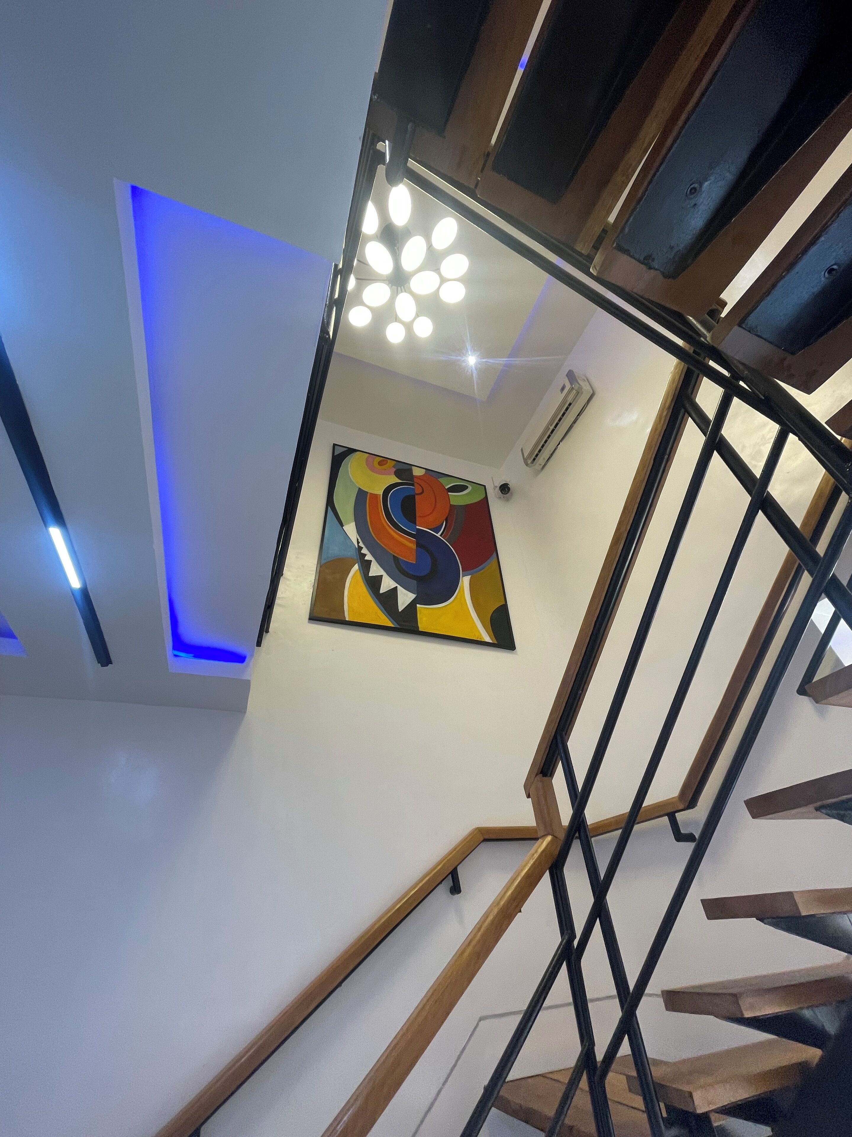 Una escalera moderna con barandilla de madera y balaustradas de metal negro, que conduce a un rellano con un cuadro abstracto de colores e iluminación empotrada.