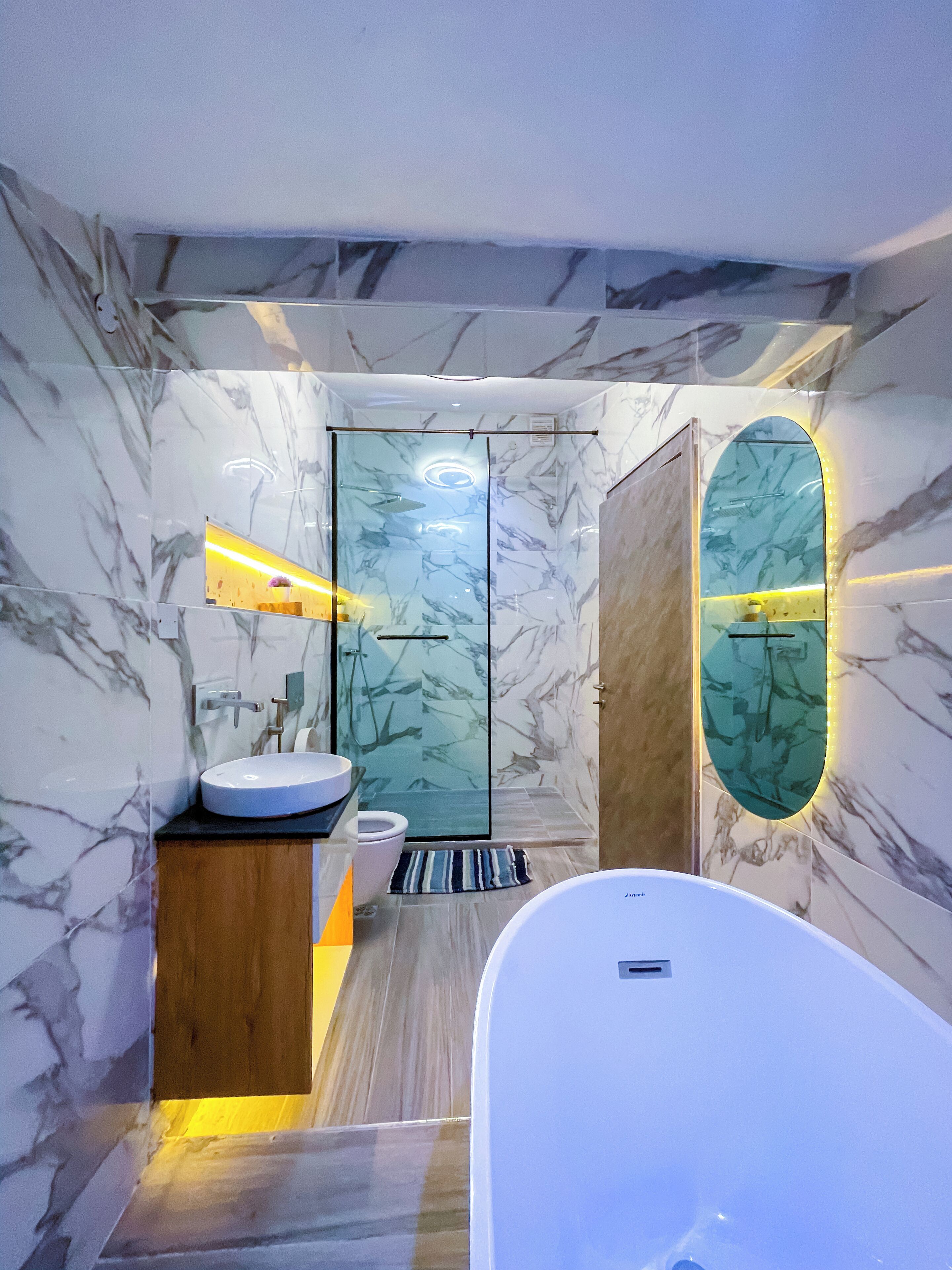 A modern bathroom with a marble wall, a wooden vanity, a white bathtub, and a glass shower enclosure.