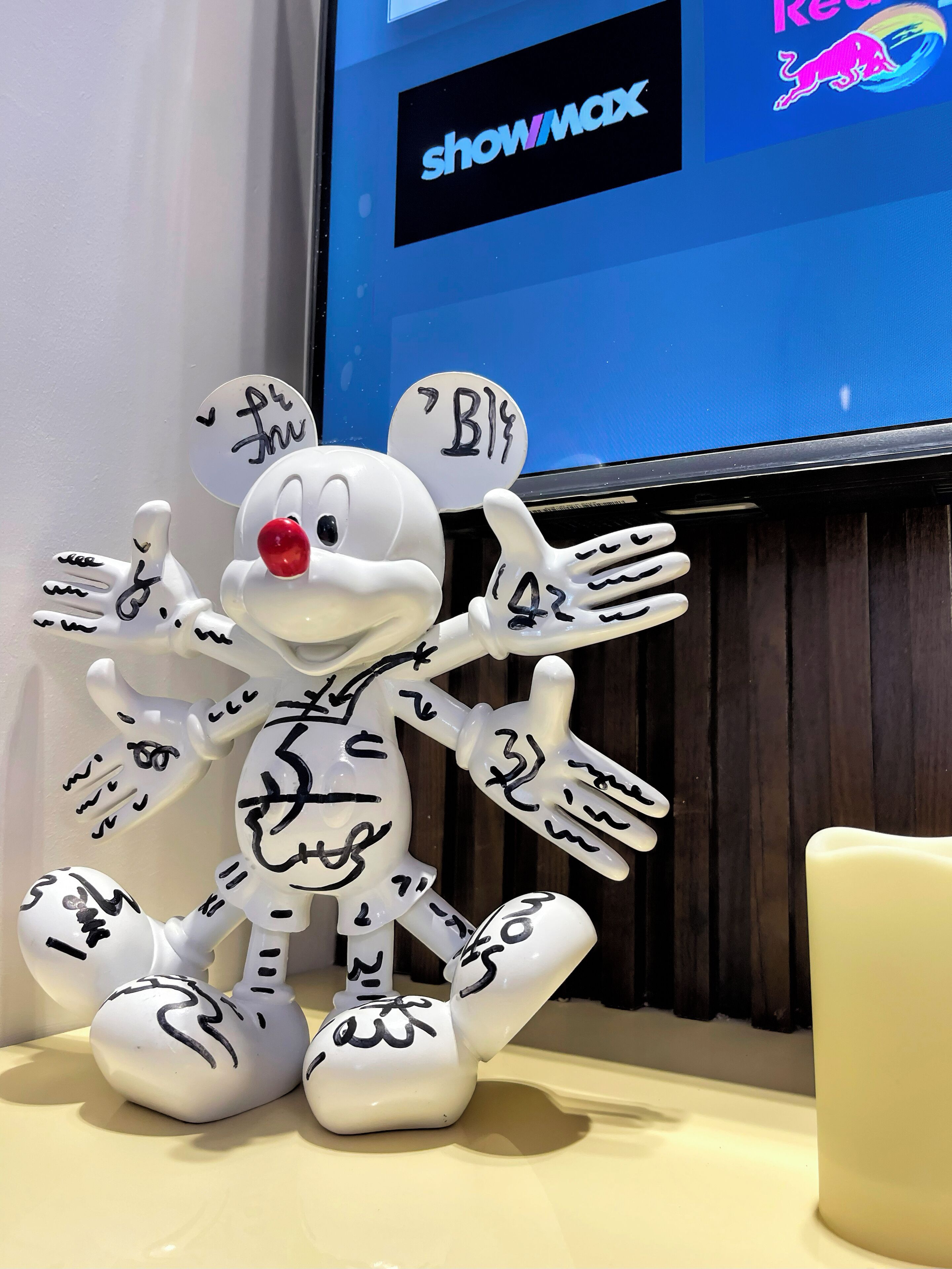 A white figurine with multiple arms and a red nose, positioned in front of a screen displaying 'showmax' and a colorful logo.
