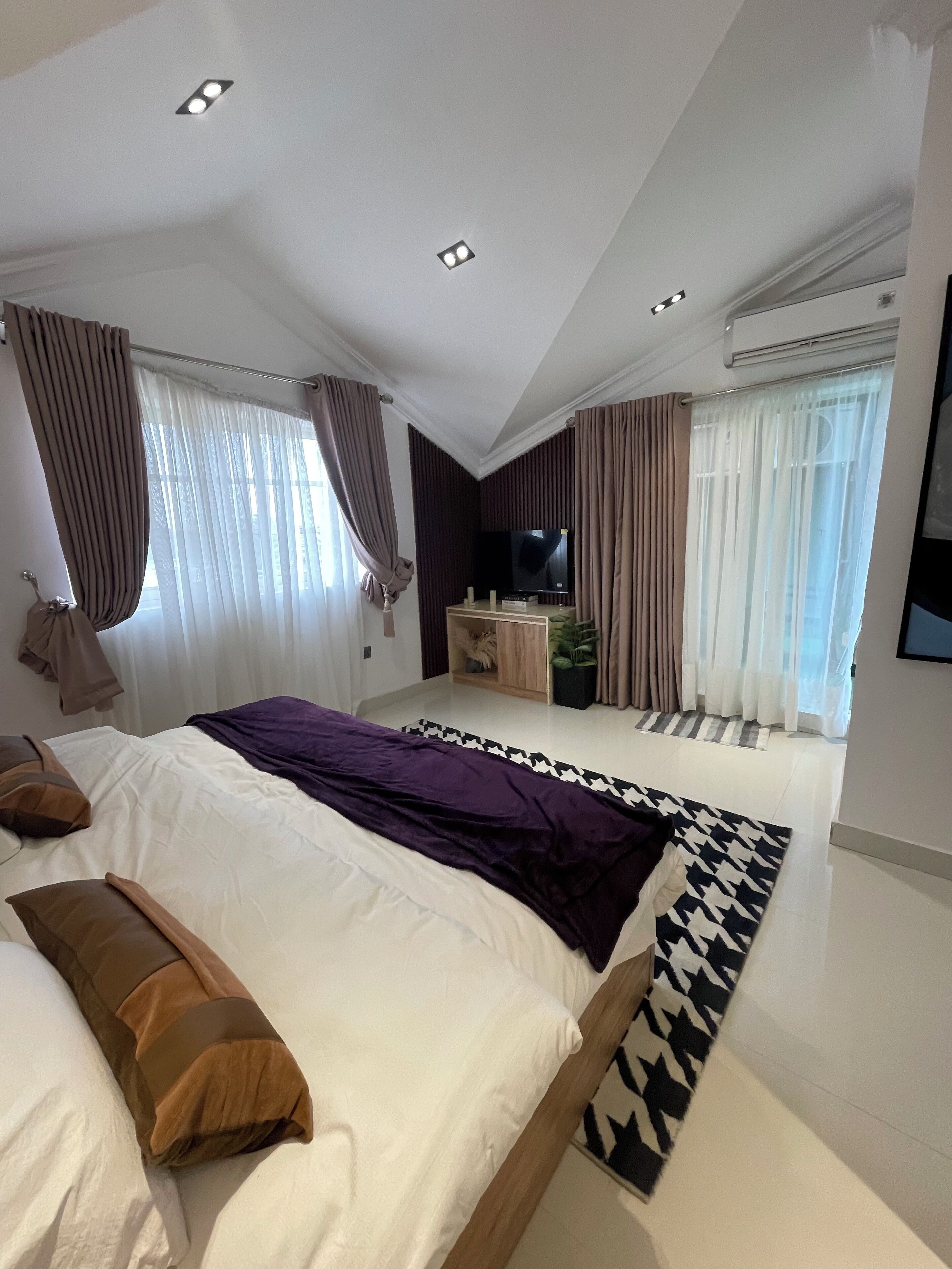 A modern bedroom with a large bed, a flat-screen TV, and a ceiling-mounted air conditioning unit.