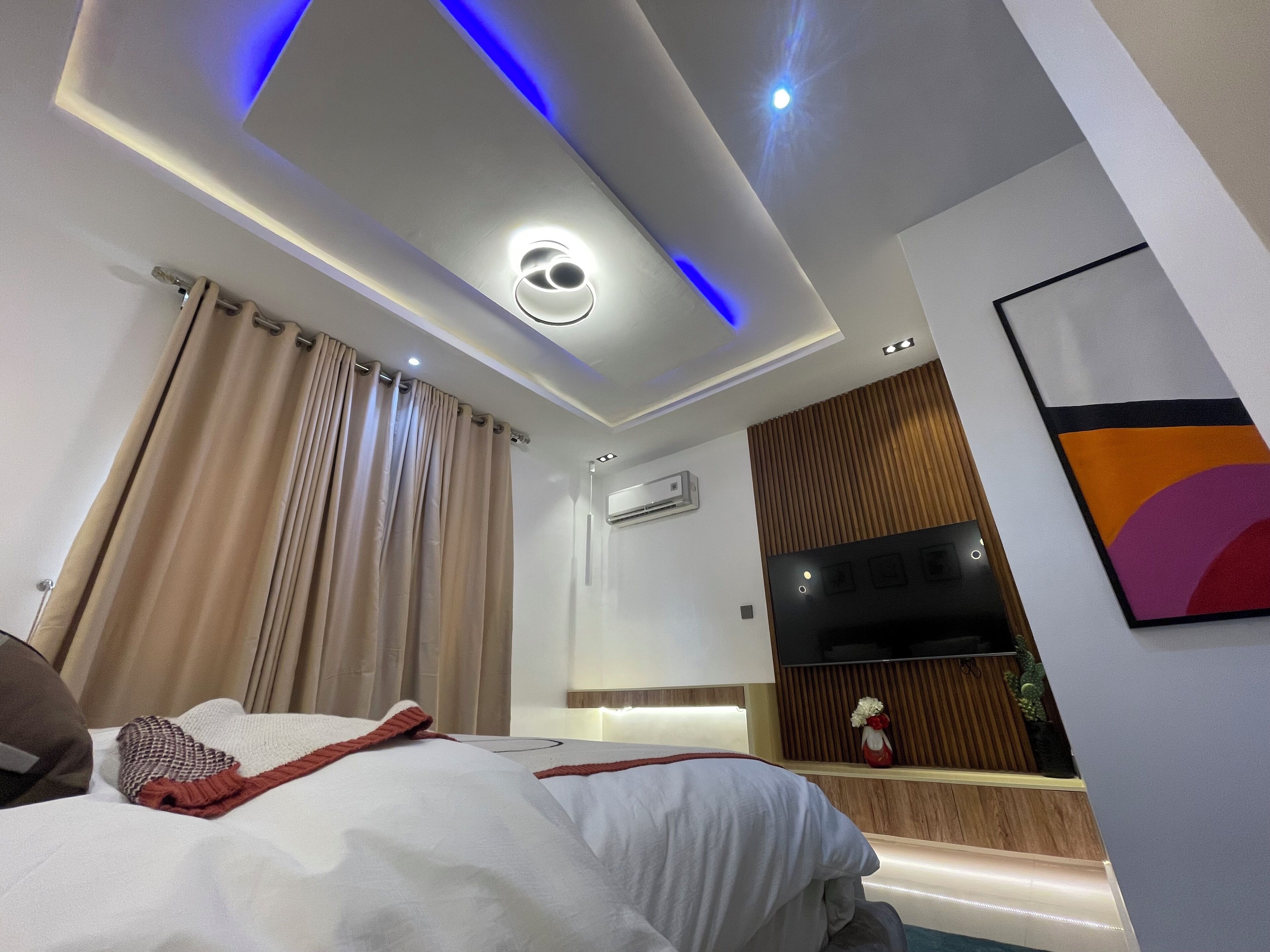 A modern hotel room with a bed, curtains, a ceiling light, an air conditioning unit, and a wall-mounted TV.