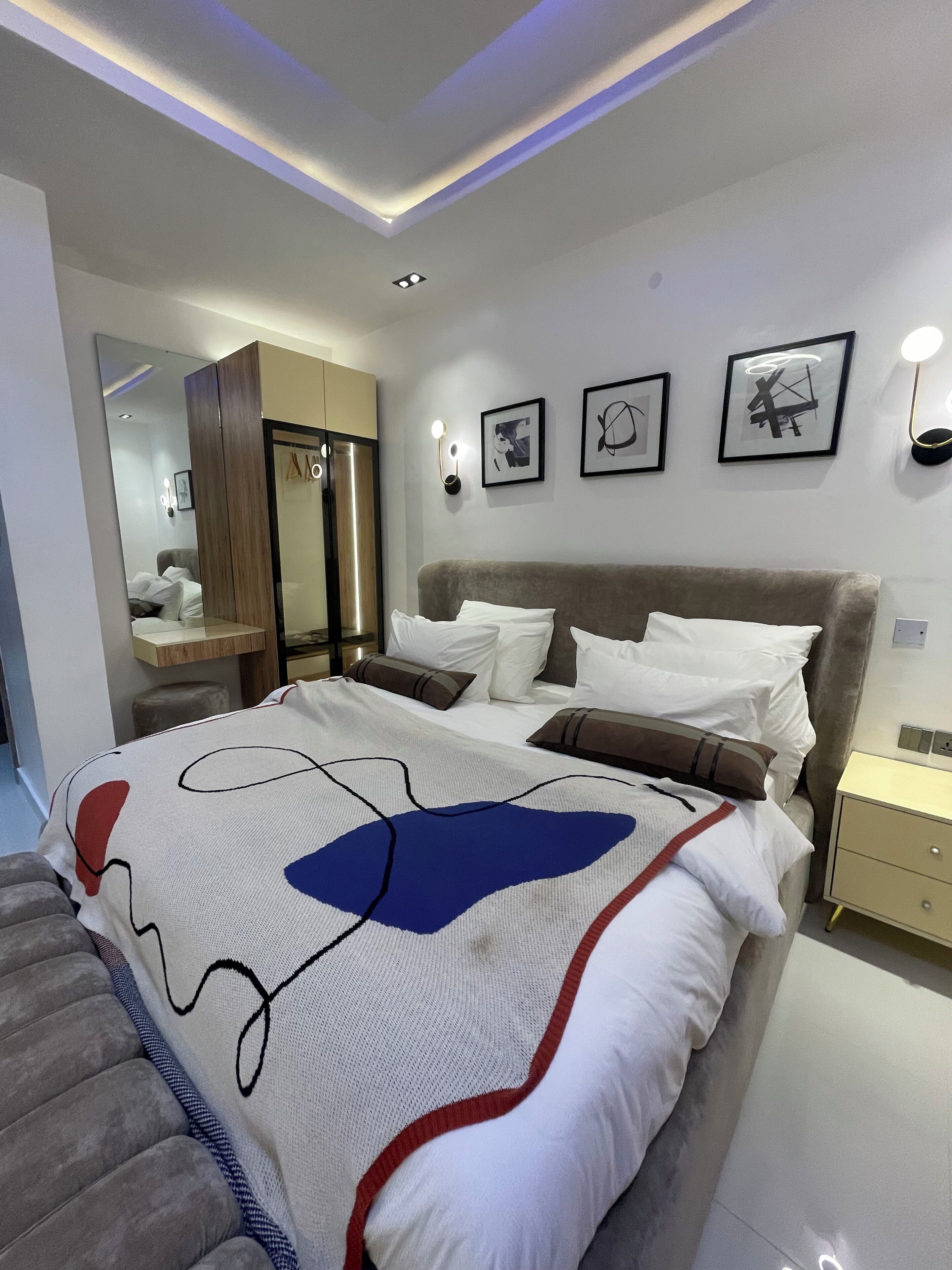 A modern bedroom with a large bed, a wardrobe, a mirror, and wall-mounted lights.