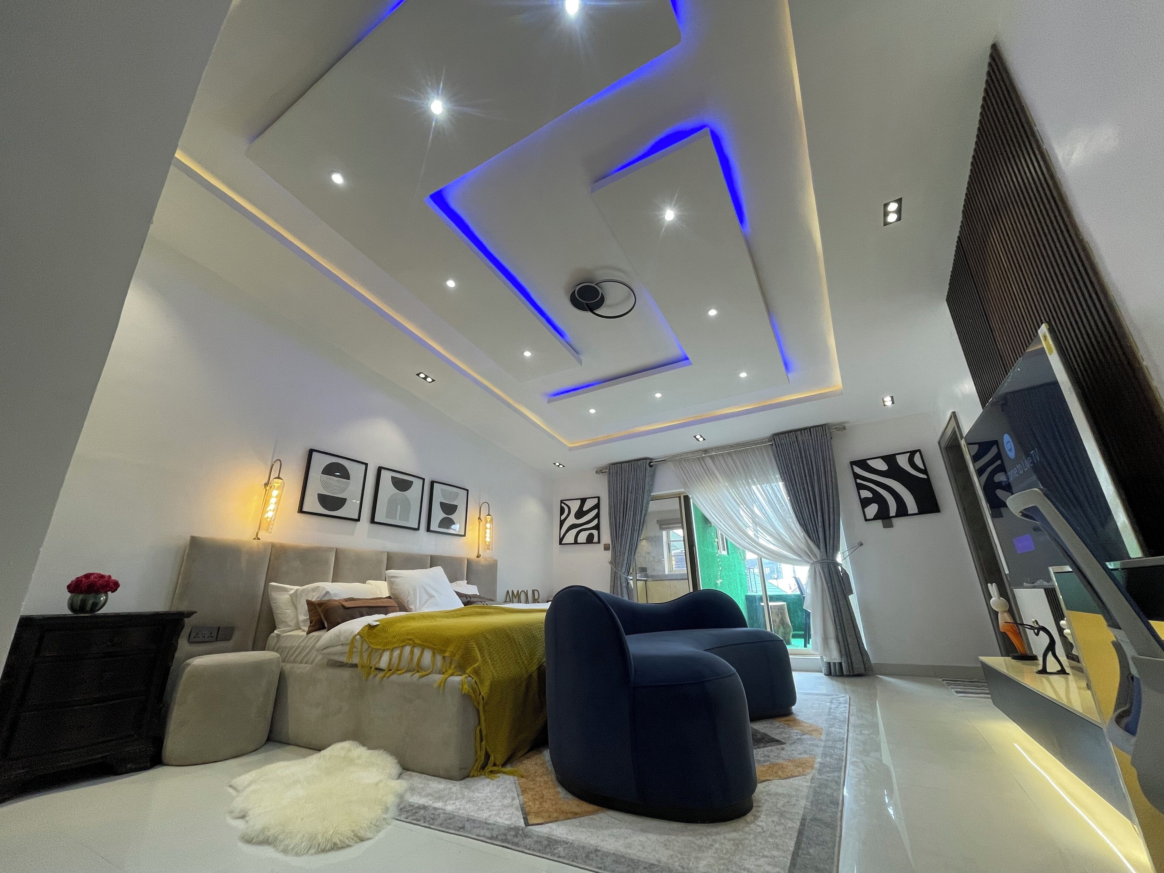 A modern bedroom with a bed, two sofas, a TV, and a ceiling with recessed lighting and blue accents.