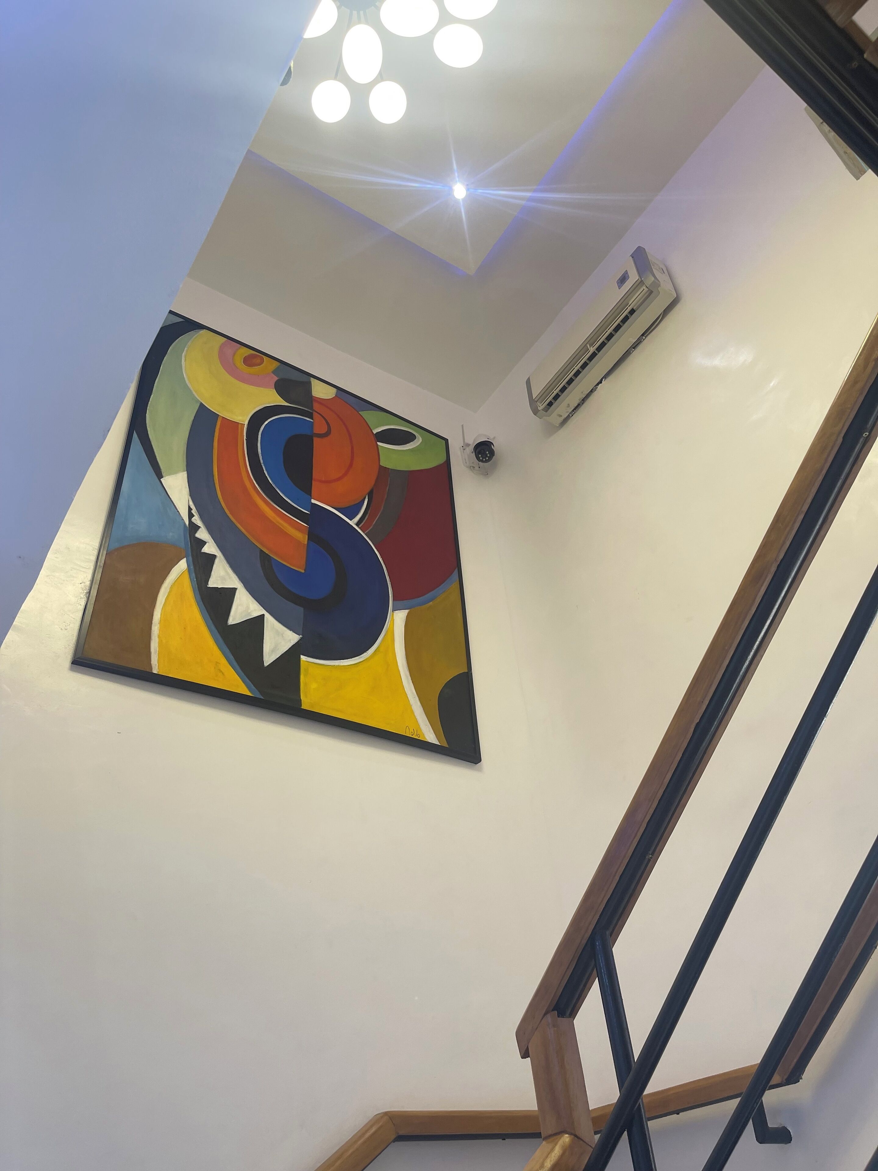A modern interior with a colorful abstract painting on the wall, a ceiling light fixture, and a staircase with wooden railings.