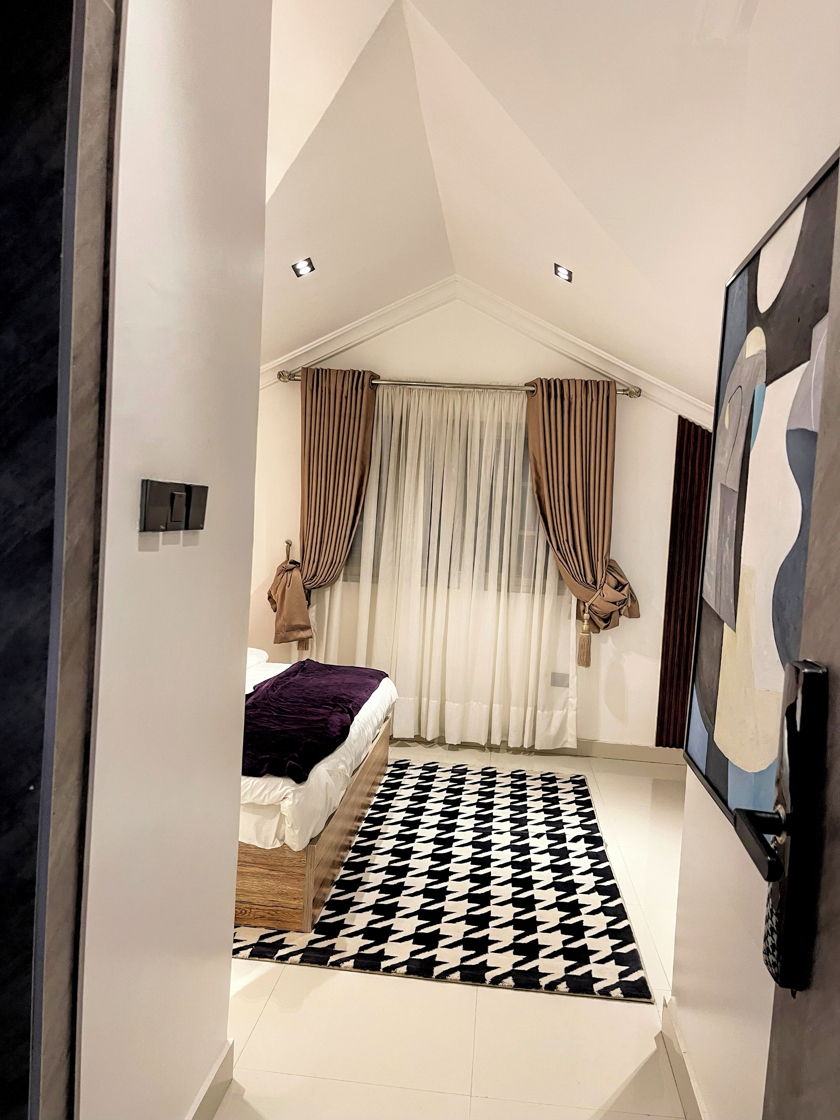 A bedroom with a bed, a patterned rug, a window with curtains, and a painting on the wall.