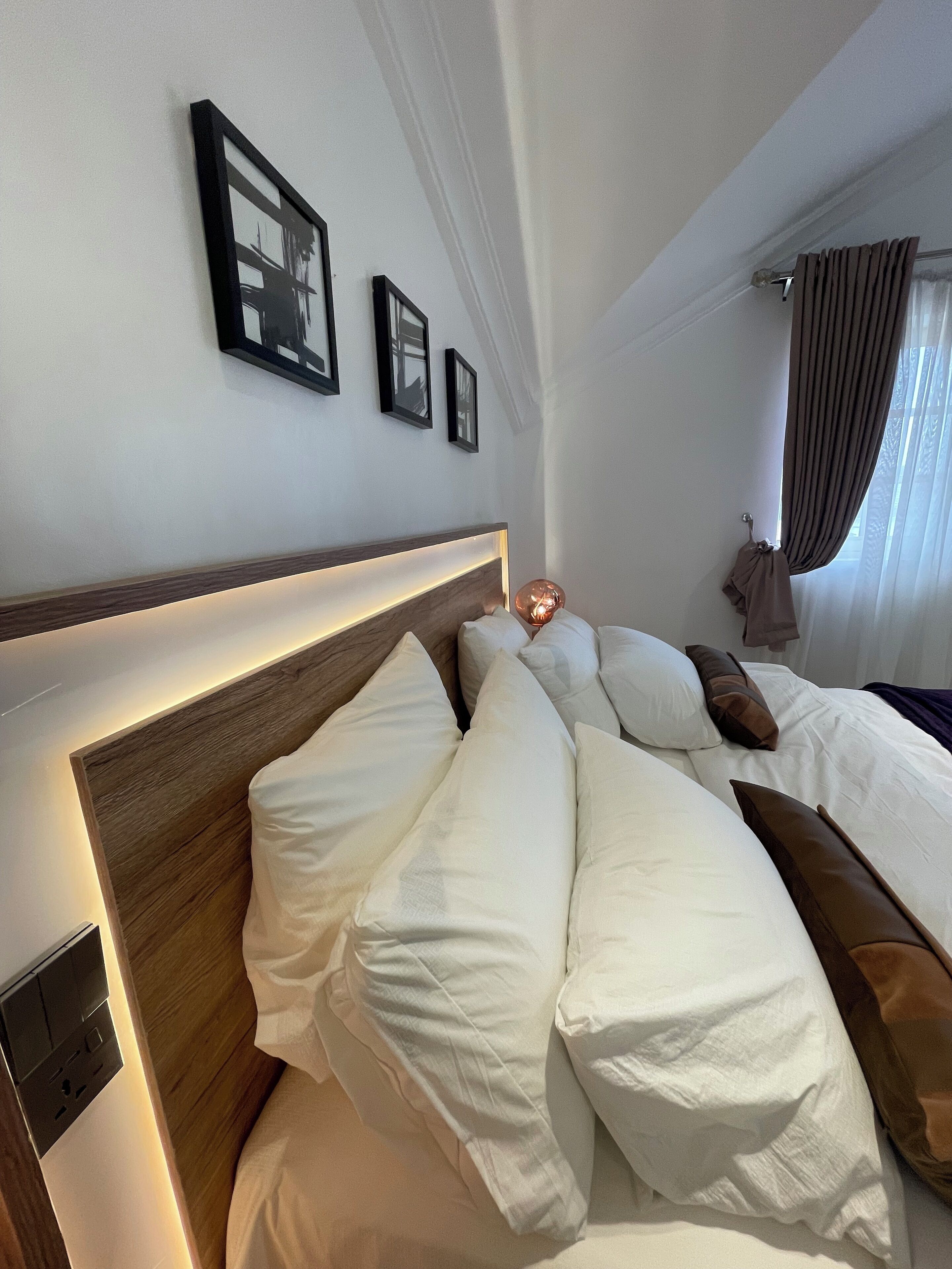 A hotel room with a neatly made bed, a headboard with integrated lighting, a bedside table, and a window with curtains.