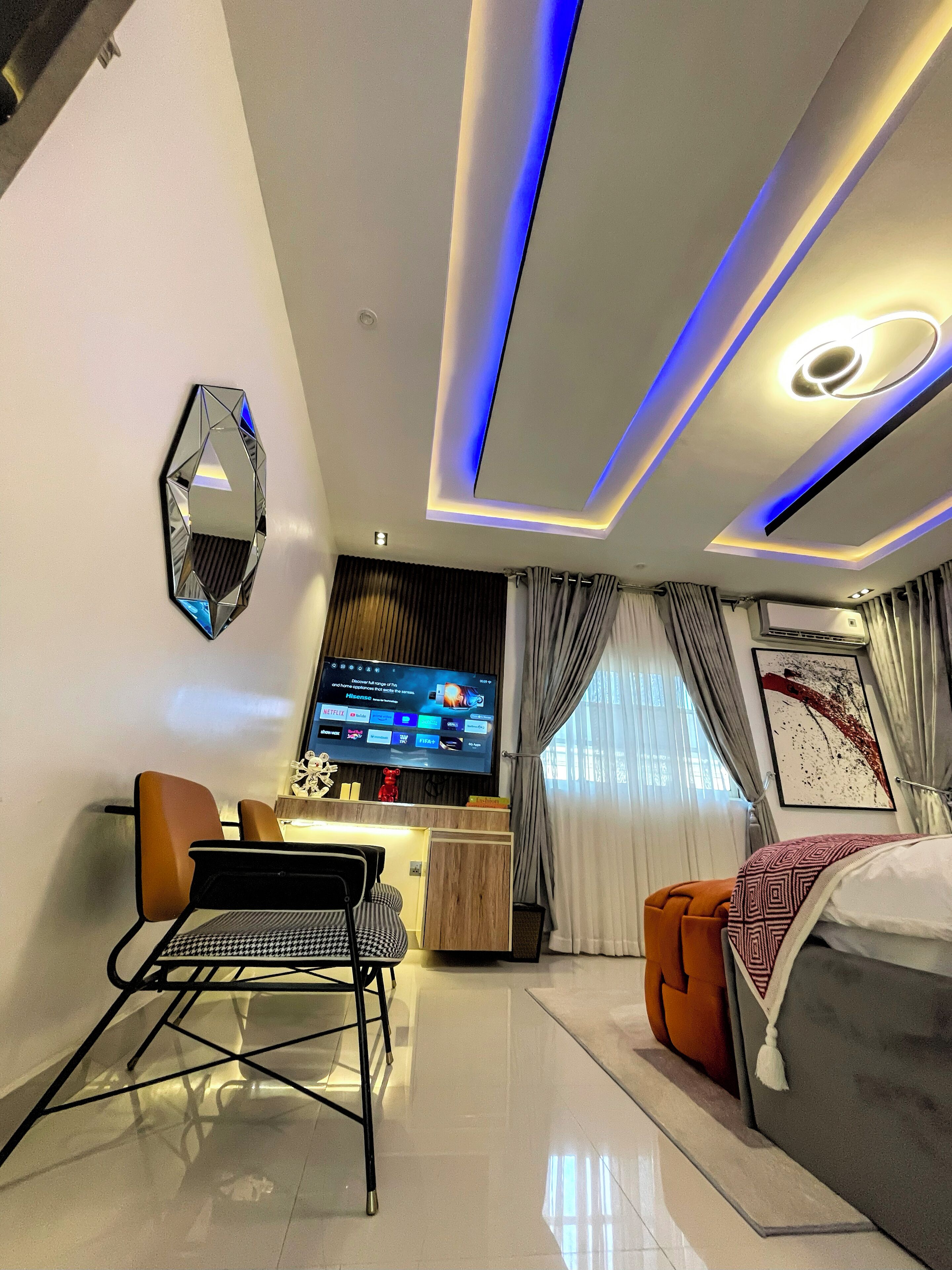 A modern hotel room with a flat-screen TV, a bed with patterned bedding, a seating area with a chair and a small table, and a decorative wall piece.