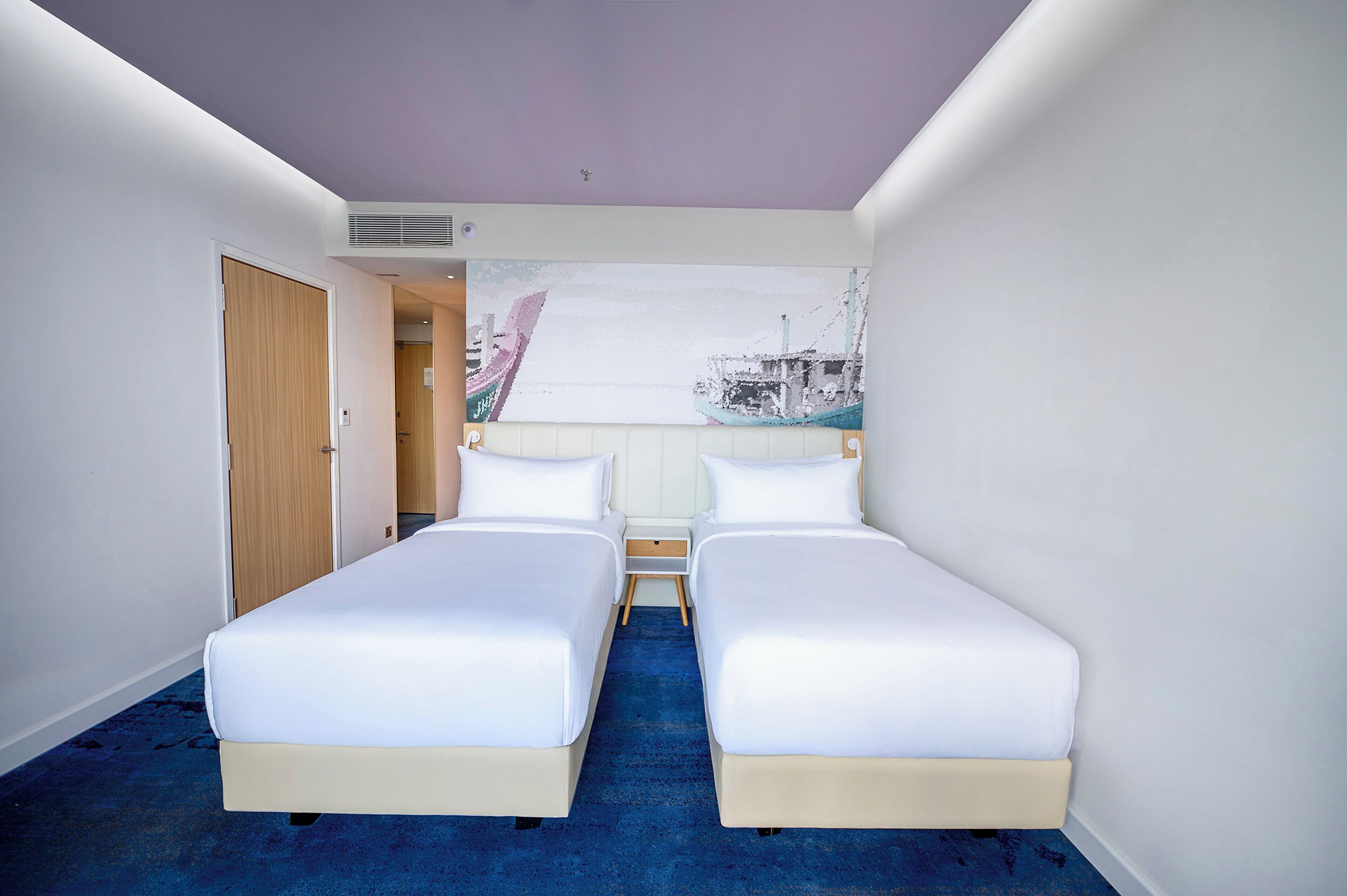 Een hotelkamer met twee enkele bedden, een houten deur, een blauw tapijt en een groot muurschilderij van een boot.