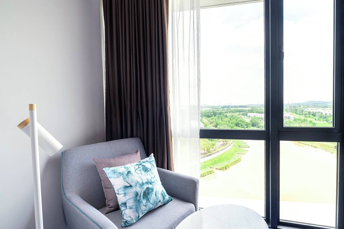 Ein modernes Zimmer mit grauem Sofa, Blick auf einen Fluss und Grünflächen, und einem großen Fenster mit Vorhängen.