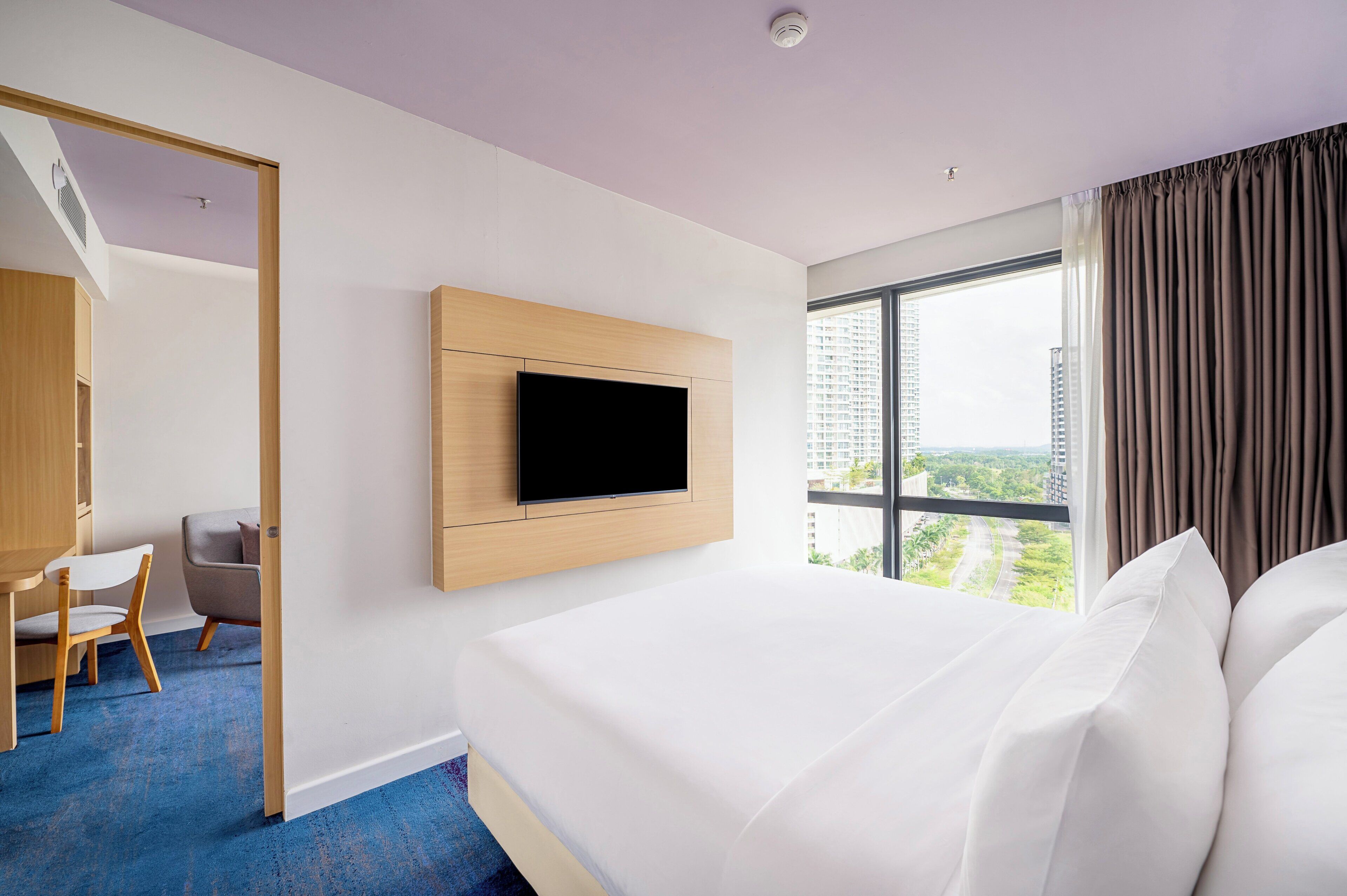 Ett hotellrum med en stor säng, en TV som hänger på väggen, ett skrivbord med en stol och utsikt över stadsbilden genom fönstret.