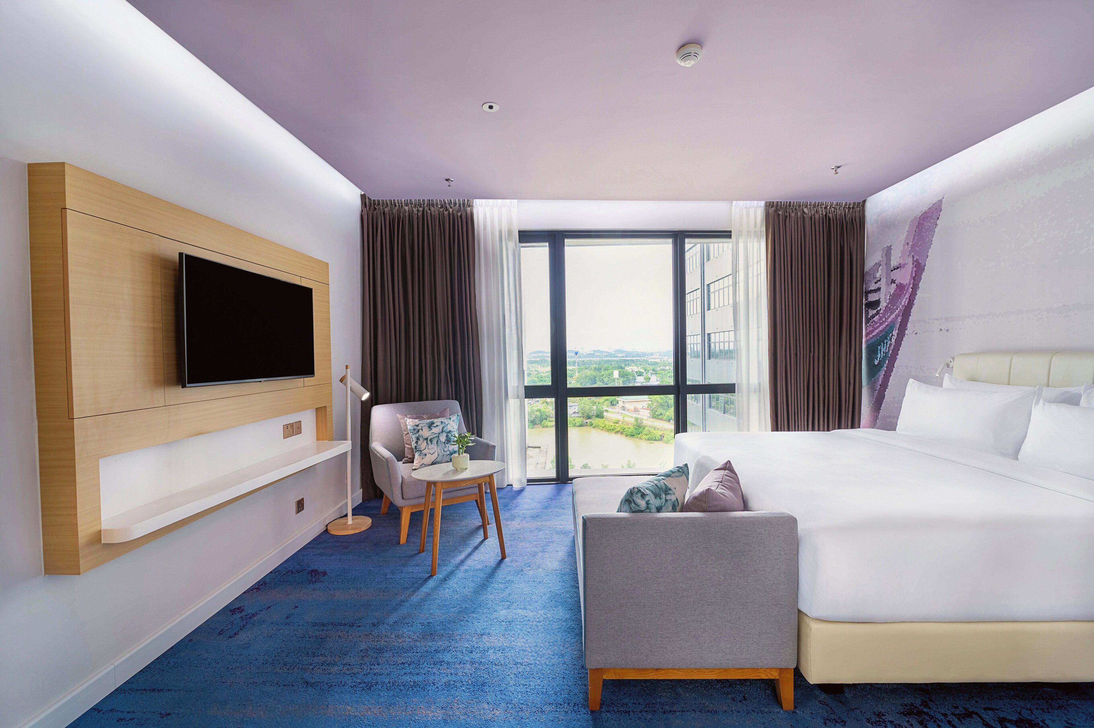 A modern hotel room with a large bed, a TV mounted on the wall, a small table with a chair, and a view of the city through large windows.