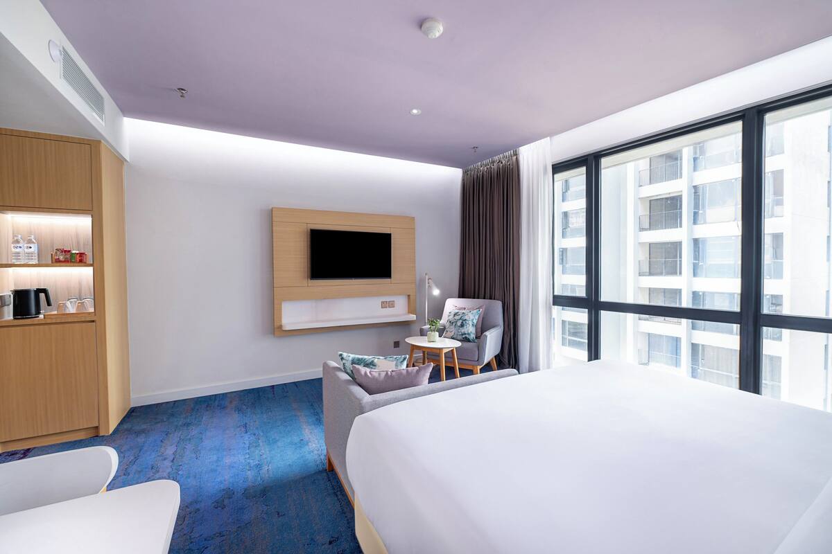 Une chambre d’hôtel avec un grand lit, un téléviseur fixé au mur, une petite table avec une chaise, et une vue sur la ville grâce à de grandes fenêtres.