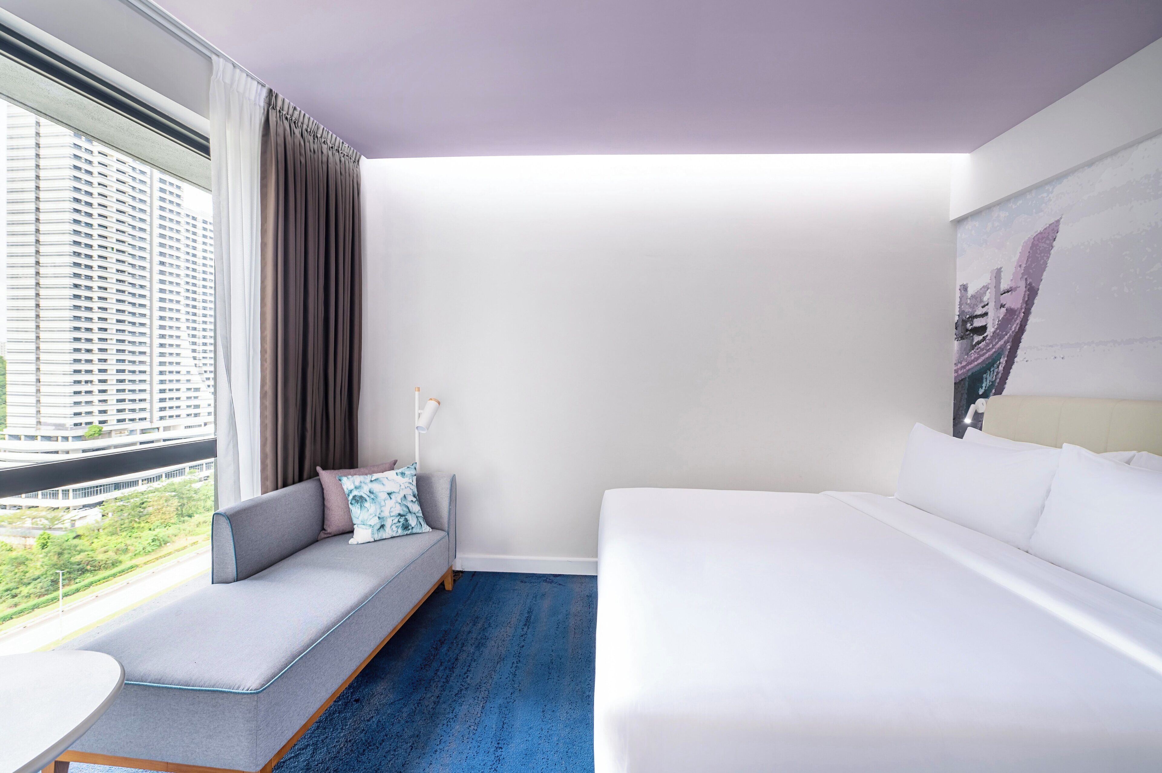 A modern hotel room with a large bed, a sofa with a view, and a painting on the wall.
