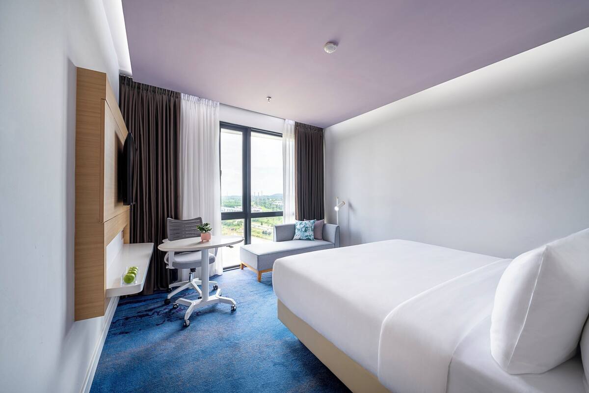 Une chambre d’hôtel moderne avec un grand lit, un bureau avec une chaise, une petite table avec une plante et une fenêtre avec des rideaux.