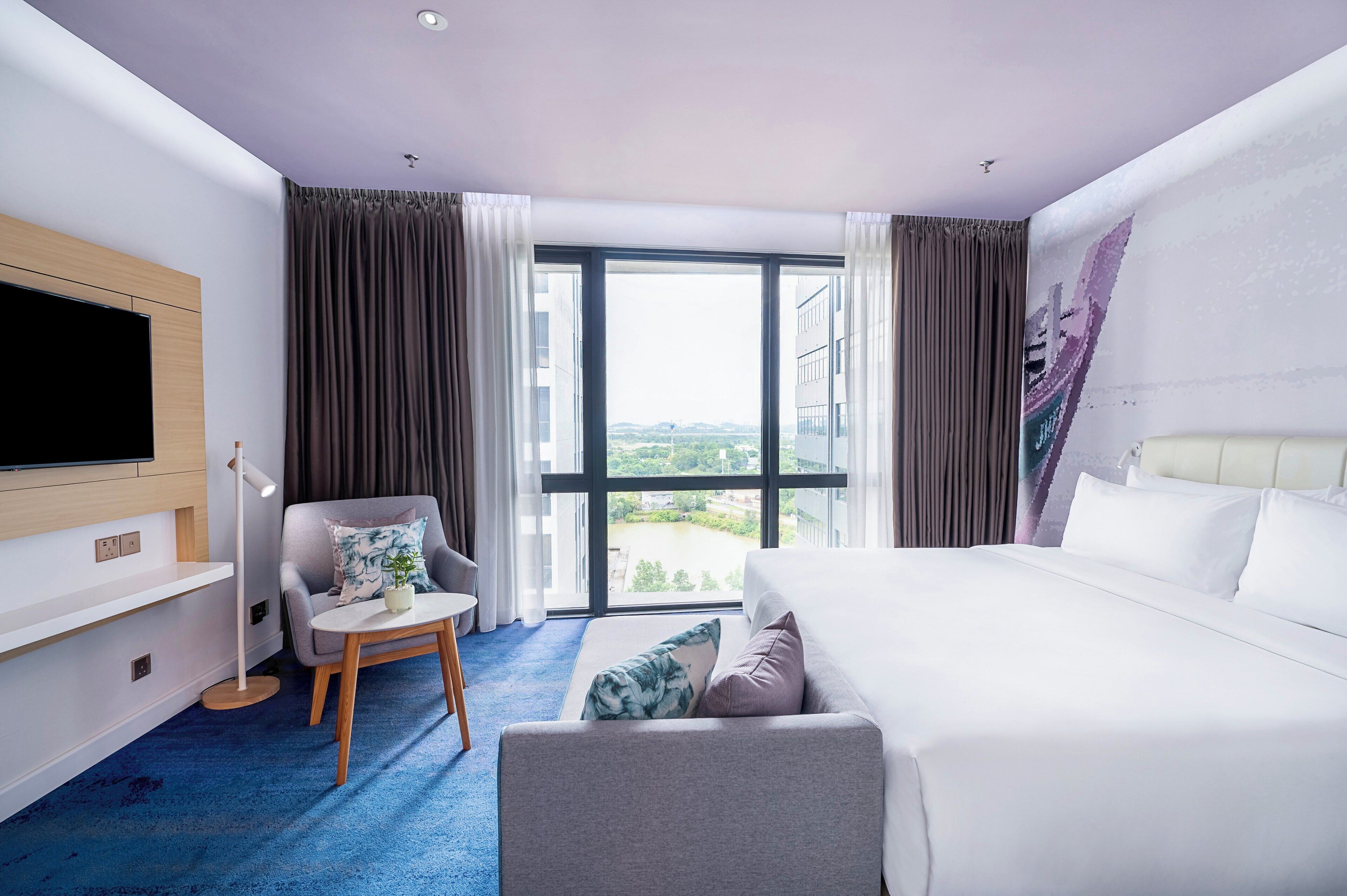 A hotel room with a large bed, a TV mounted on the wall, a small table with a chair, and a view of the city through large windows.