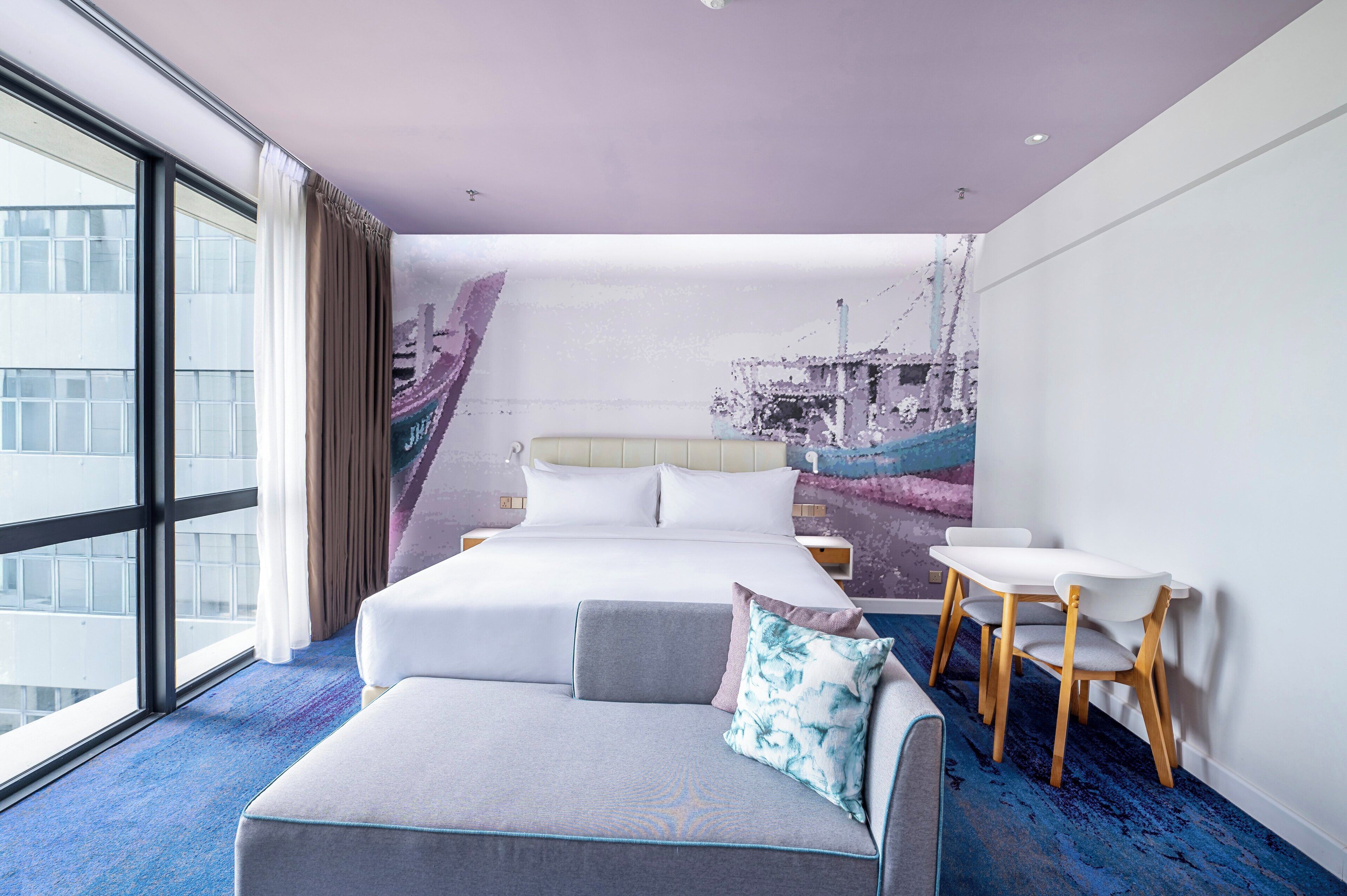 A hotel room with a large bed, a sofa, a small table, and a view of the city through large windows.