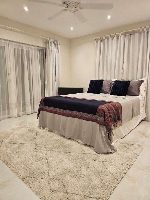 Un dormitorio con cama, mesita de noche, ventilador de techo y cortinas traslúcidas.