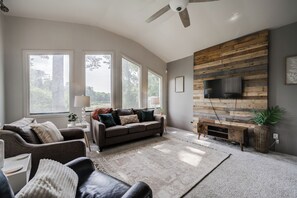 A spacious living room with a large window, a ceiling fan, a wooden wall panel, and a flat-screen TV mounted on the wall. - Pinnacle Point~ 2min to Pinnacle Mtn, Deck, Pond View, Spacious (Little Rock)