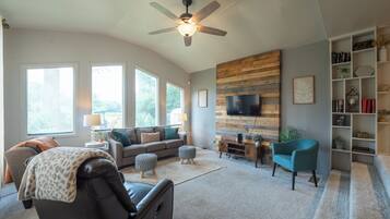 A spacious living room with a ceiling fan, a large TV mounted on a wooden wall, a sofa set, ottomans, and a bookshelf.