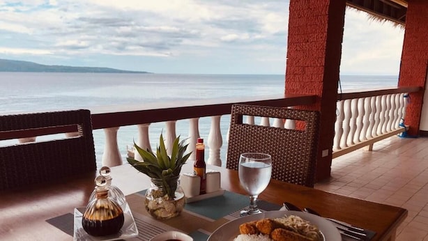 Una mesa de comedor con comida, un vaso de agua y condimentos en un balcón con vista al mar.