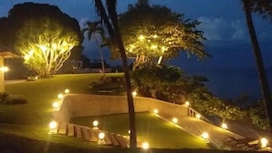 Un área ajardinada con senderos, árboles iluminados y una piscina pequeña, con vista al mar.